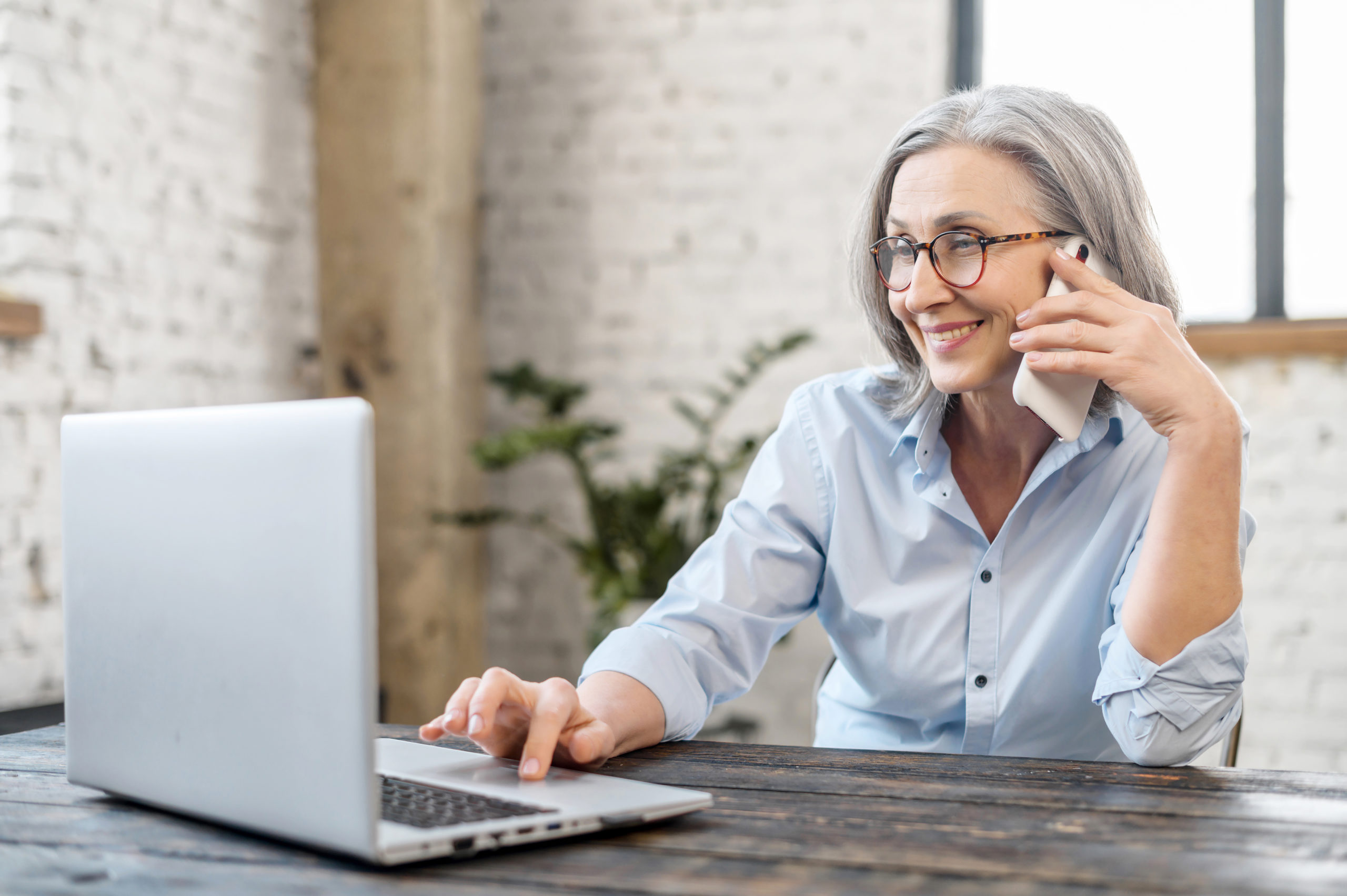 Senior bossy businesswoman has phone conversation working with a laptop at the desk in office. An aged saleswoman talking with a customers by smartphone. Smiling old woman speaking on the smartphone